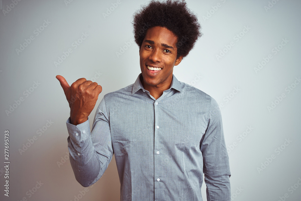 Young african american man with afro hair wearing grey shirt over isolated white background smiling with happy face looking and pointing to the side with thumb up. Young african american man with afro hair wearing grey shirt over isolated white background smiling with happy face looking and pointing to the side with thumb up.