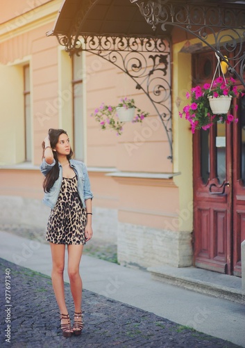 Stylish long hair brunette woman in a leopard animal print dress with sexy legs in brown high heel shoes, denim jacket, street fashion concept. Fashionable girl walking on summer street. Trendy style