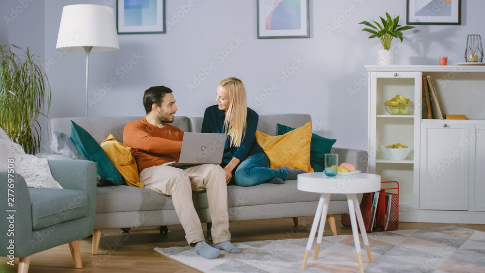 Happy Lovely Couple Sits on a Sofa at Home, Boyfriend Holds Laptop on Knees, Browsing through Internet, Doing e-Shopping, Using Streaming Services. Happy Family in Cozy Living Room. Happy Lovely Couple Sits on a Sofa at Home, Boyfriend Holds Laptop on Knees, Browsing through Internet, Doing e-Shopping, Using Streaming Services. Happy Family in Cozy Living Room.