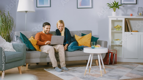 Happy Lovely Couple Sits on a Sofa at Home, Boyfriend Holds Laptop on Knees, Browsing through Internet, Doing e-Shopping, Using Streaming Services. Happy Family in Cozy Living Room. © Gorodenkoff