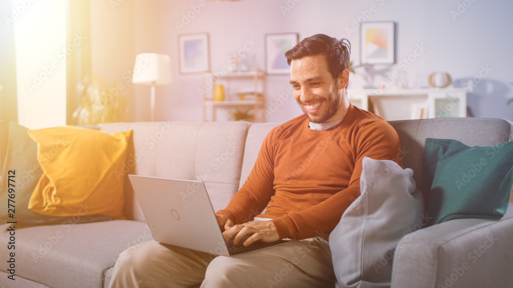 Cheerful Young Man Sitting on a Sofa Holds Laptop on His Lap, Browses Through the Internet, Social Networks, Does e-Shopping. Man at Home Using Laptop while Sitting on a Couch. Warm Sun Light. Cheerful Young Man Sitting on a Sofa Holds Laptop on His Lap, Browses Through the Internet, Social Networks, Does e-Shopping. Man at Home Using Laptop while Sitting on a Couch. Warm Sun Light.