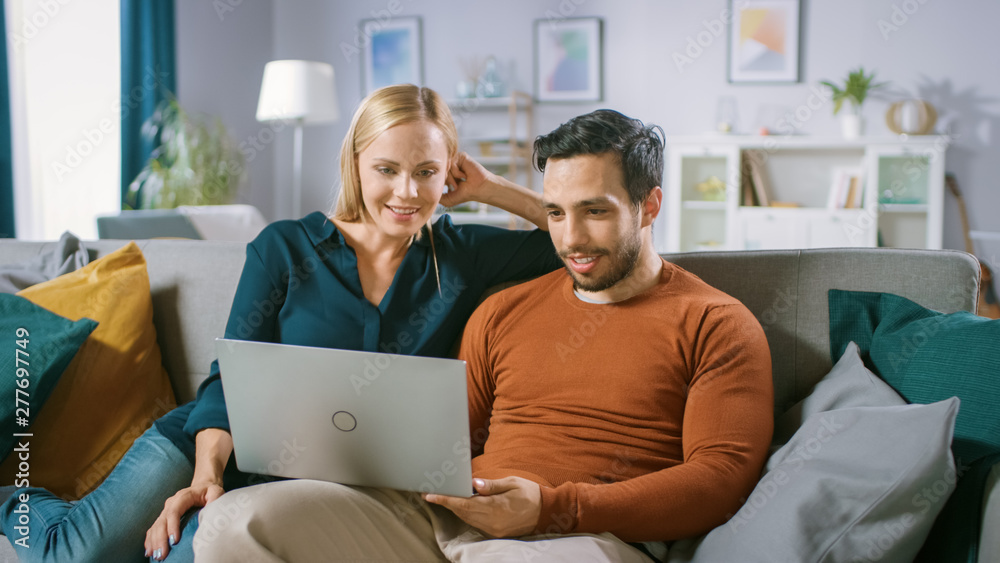 Happy Lovely Couple Sits on a Sofa at Home, Boyfriend Holds, Browsing through Internet, Doing e-Shopping, Using Streaming Services. Happy Family in Cozy Living Room. Happy Lovely Couple Sits on a Sofa at Home, Boyfriend Holds, Browsing through Internet, Doing e-Shopping, Using Streaming Services. Happy Family in Cozy Living Room.
