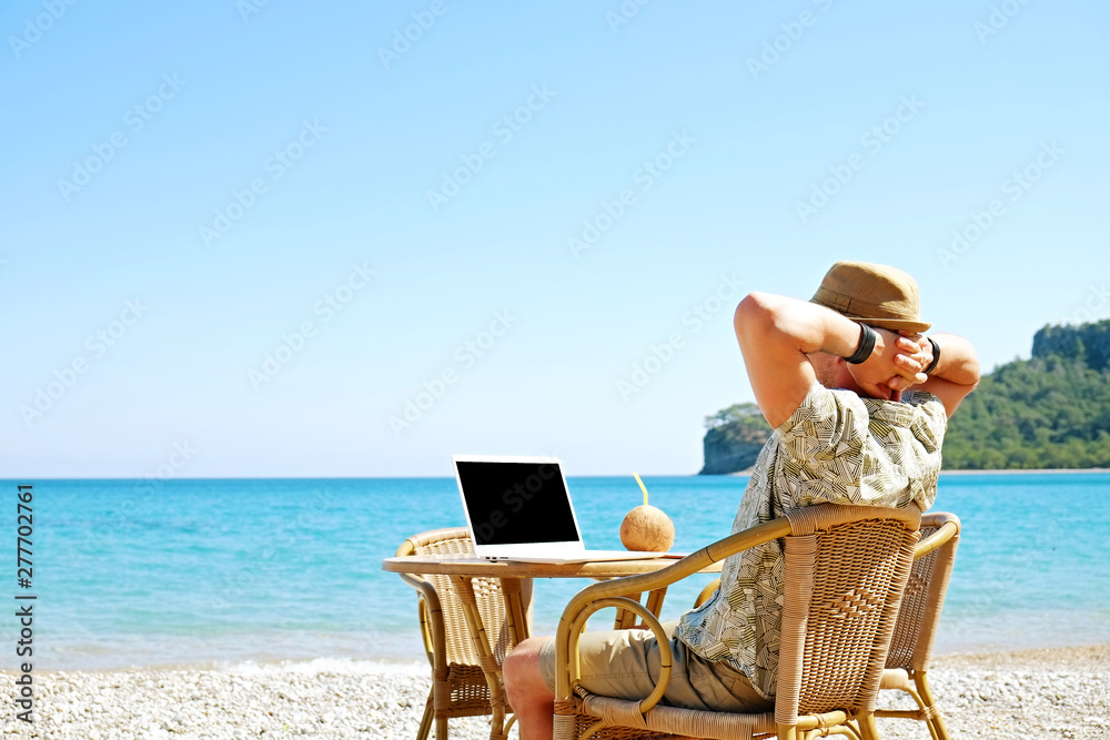 Fit travel blogger sitting at beach bar writing article on white laptop. Freelance remote work concept. Self employed man coding, wearing typical tourist shirt and hat. Copy space, sea view background