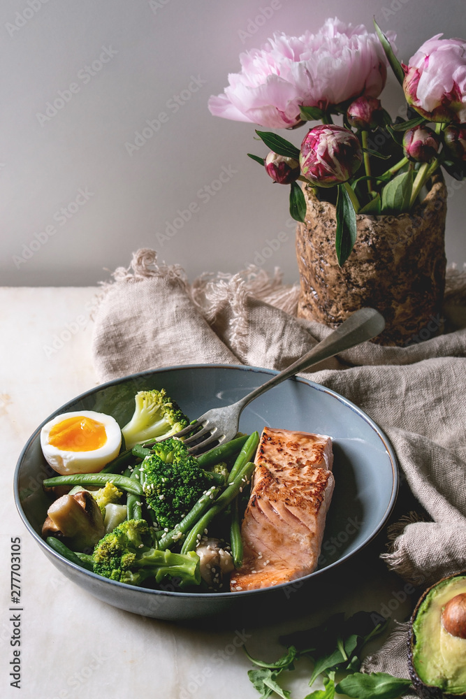 Ketogenic low carb diet dinner grilled salmon, avocado, broccoli, green bean and soft boiled egg in ceramic bowl served with olives and nuts, bouquet of peonies on grey table. - obrazy, fototapety, plakaty 