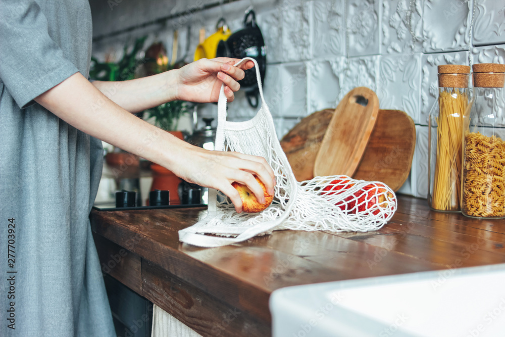Young woman in grey dress pulls apples out of knitted rag bag string bag shopper in the kitchen, zero waste, slow life - obrazy, fototapety, plakaty 