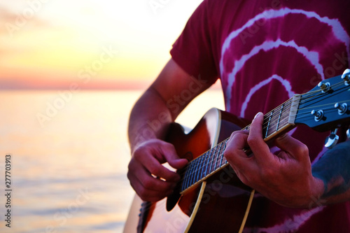 Young man wearing purple tie dye t-shirt playing dreadnought parlor acoustic guitar on beach at beautiful sunset time. Fit guitarist w/ sunburst instrument by the sea. Background, copy space, close up photo