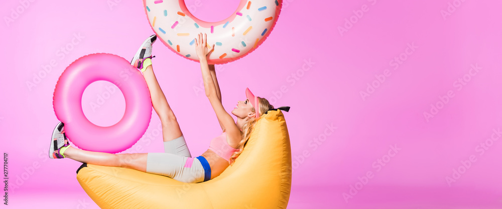 panoramic shot of girl on bean bag chair with inflatable swim rings on pink, doll concept - obrazy, fototapety, plakaty 