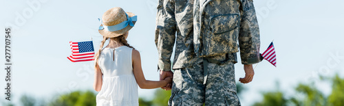 Panoramic shot of patriotic child and man in military uniform holding hands and American flags
