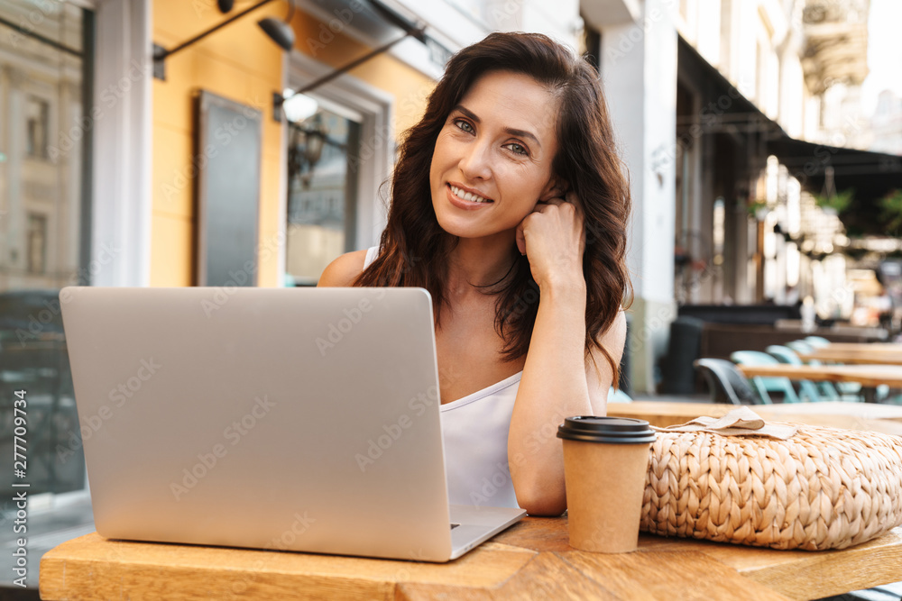 Portrait of smiling seductive woman drinking coffee from paper cup and using laptop while sitting in cozy cafe outdoors Portrait of smiling seductive woman drinking coffee from paper cup and using laptop while sitting in cozy cafe outdoors