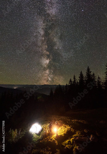 Vertical shot, top view of summer camping in mountains. Young couple hikers standing together beside campfire and tourist tent. On background beautiful night starry sky full of stars and Milky way.