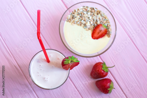 A glass of strawberry yogurt, a deep plate of muesli with strawberries stand on a delicate pink wooden background. Top view on the center of the frame.