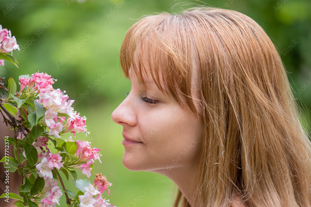 Beautiful young woman sniffing flowers with her eyes closed and smiling. Close up portrait of face girl with flowering branch at park outdoors. Beautiful young woman sniffing flowers with her eyes closed and smiling. Close up portrait of face girl with flowering branch at park outdoors.