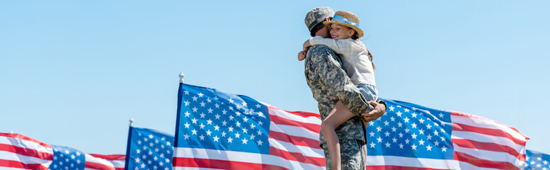 Panoramic shot of military man holding in arms cheerful child near American flags