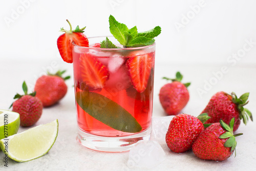 On a light background there is a strawberry cocktail in a glass with ice and mint   strawberries and lime slices .