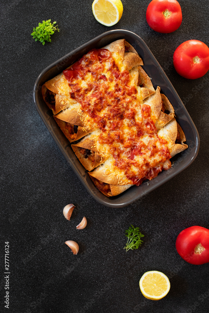 Mexican enchiladas with chicken, vegetables, corn, beans, tomato sauce and cheese. Served in baking tray. Mexican food. Latin American cuisine. Black background, top view, copy space
