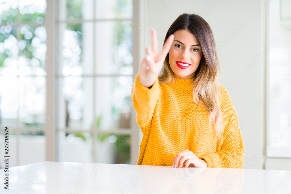 Young beautiful woman wearing winter sweater at home smiling looking to the camera showing fingers doing victory sign. Number two. Young beautiful woman wearing winter sweater at home smiling looking to the camera showing fingers doing victory sign. Number two.