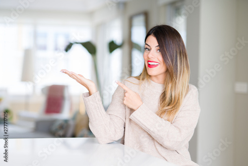 Free Download Young beautiful woman wearing winter sweater at home amazed and smiling to the camera while presenting with hand and pointing with finger. - GFXLoad