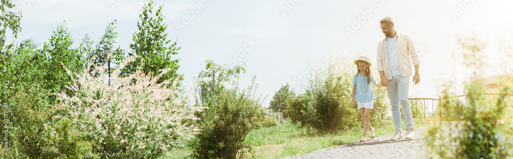 panoramic shot of handsome father and daughter walking near green plants and holding hands