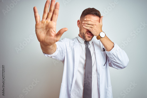 Young handsome doctor man wearing white profressional coat over isolated background covering eyes with hands and doing stop gesture with sad and fear expression. Embarrassed and negative concept. photo