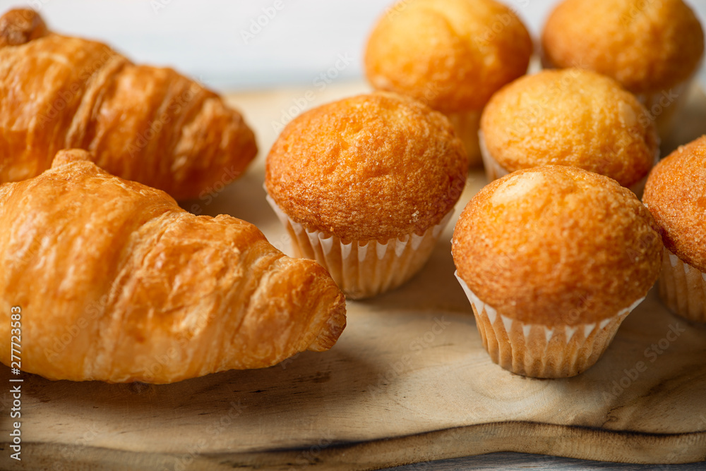 Close-up of cupcakes and croissants prepared for breakfast on wooden board on white wooden table. Close-up of cupcakes and croissants prepared for breakfast on wooden board on white wooden table.