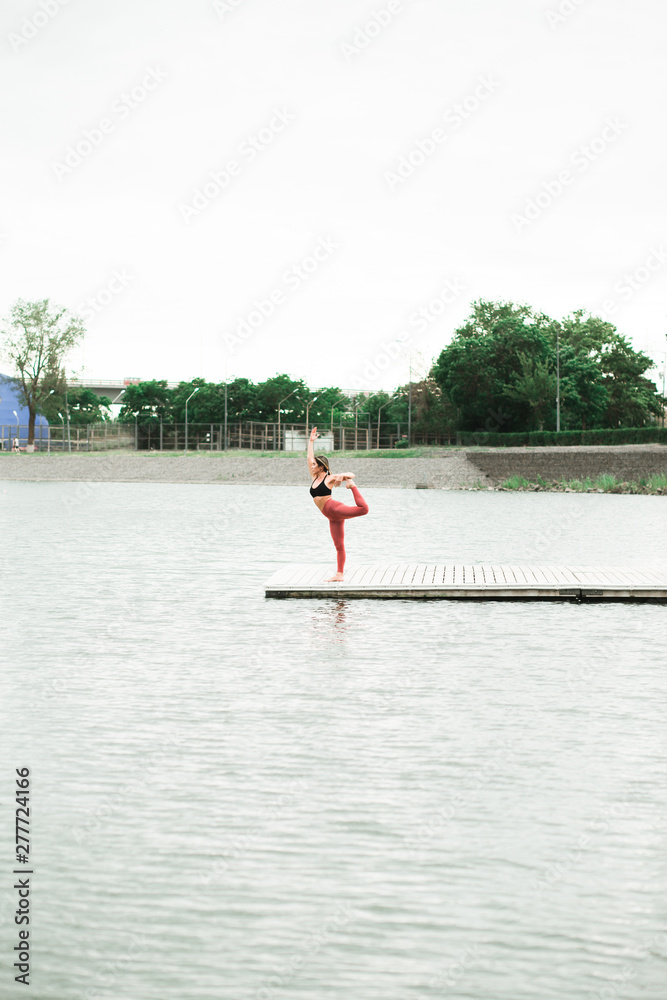 Fototapeta premium Asian young girl doing yoga outdoors on the pier by the lake.