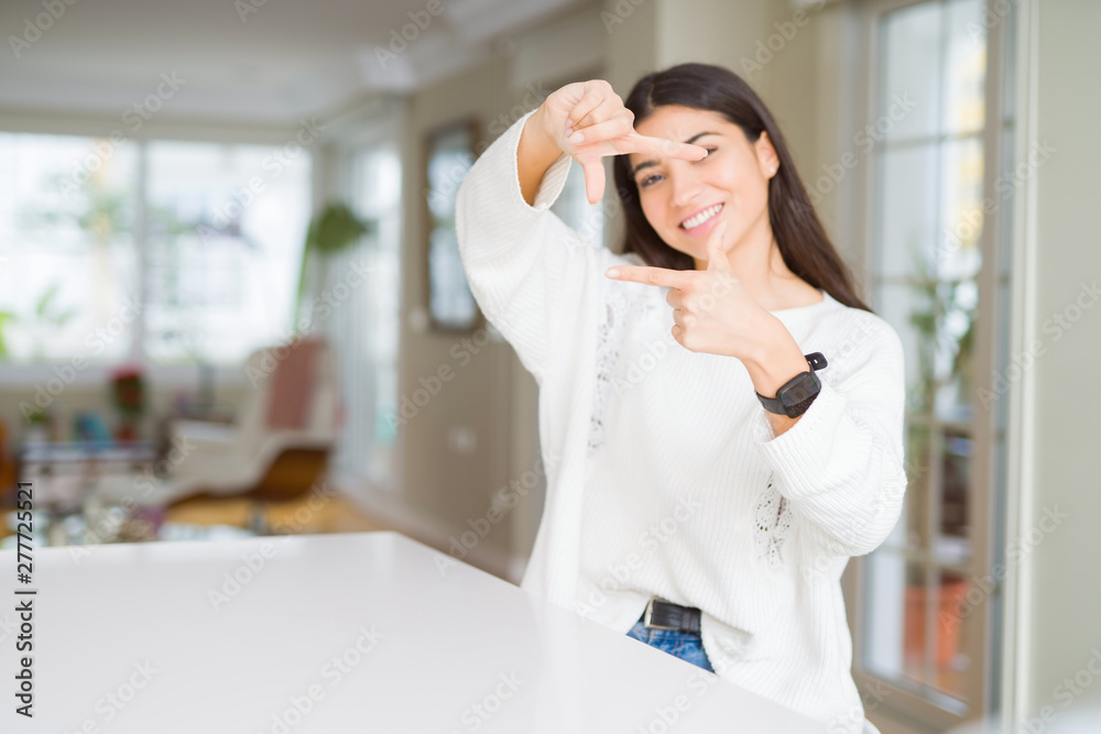 Young beautiful woman at home on white table smiling making frame with hands and fingers with happy face. Creativity and photography concept. Young beautiful woman at home on white table smiling making frame with hands and fingers with happy face. Creativity and photography concept.