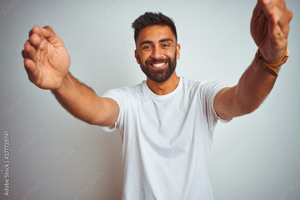 Young indian man wearing t-shirt standing over isolated white background looking at the camera smiling with open arms for hug. Cheerful expression embracing happiness. Young indian man wearing t-shirt standing over isolated white background looking at the camera smiling with open arms for hug. Cheerful expression embracing happiness.