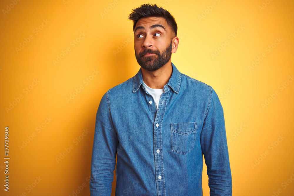 Young indian man wearing denim shirt standing over isolated yellow background smiling looking to the side and staring away thinking. Young indian man wearing denim shirt standing over isolated yellow background smiling looking to the side and staring away thinking.