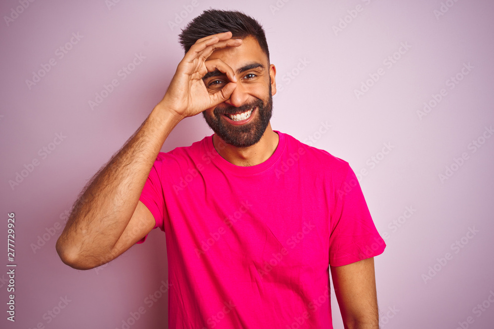 Young indian man wearing t-shirt standing over isolated pink background doing ok gesture with hand smiling, eye looking through fingers with happy face. Young indian man wearing t-shirt standing over isolated pink background doing ok gesture with hand smiling, eye looking through fingers with happy face.