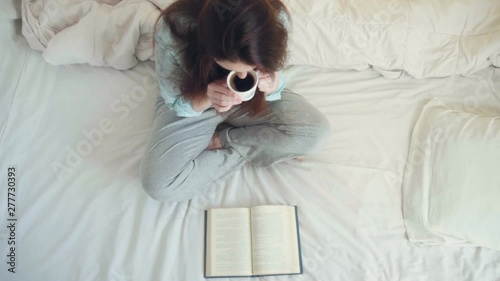 Beautiful woman enjoying drinking warm coffee and reading book on bed