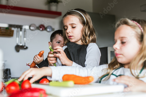 Little girls and boy cutting and peeling ripe vegetables while cooking healthy salad in kitchen together