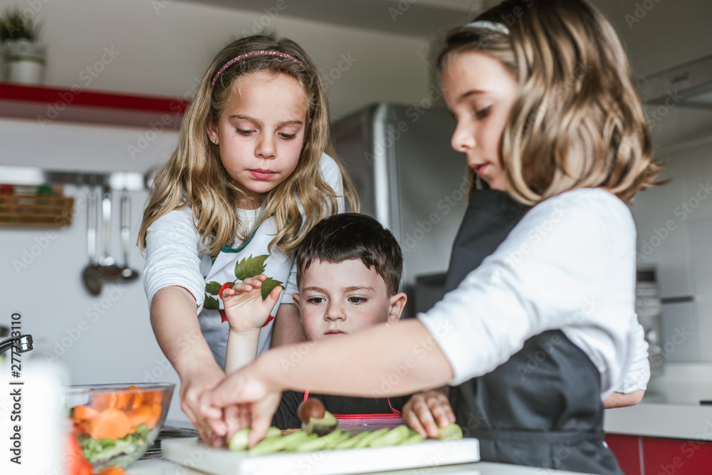 Little girls and boy cutting and peeling ripe vegetables while cooking healthy salad in kitchen together Little girls and boy cutting and peeling ripe vegetables while cooking healthy salad in kitchen together