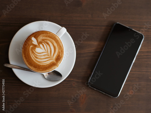 Overhead smartphone and fresh cappuccino coffee in white ceramic cup, saucer, spoon with beautiful handmade latte art on wooden table in cozy city cafe ready to drink