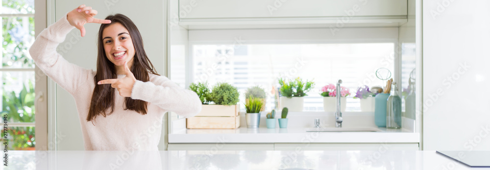 Wide angle picture of beautiful young woman sitting on white table at home smiling making frame with hands and fingers with happy face. Creativity and photography concept. Wide angle picture of beautiful young woman sitting on white table at home smiling making frame with hands and fingers with happy face. Creativity and photography concept.