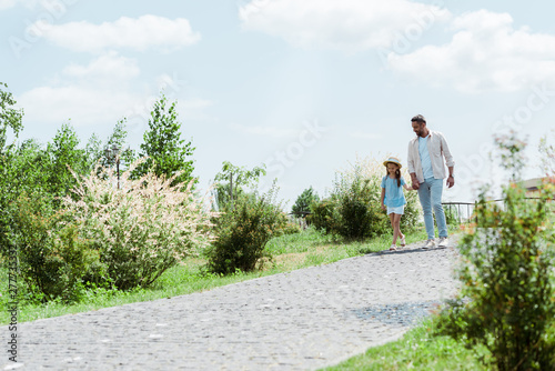 handsome father and daughter walking near green plants and holding hands