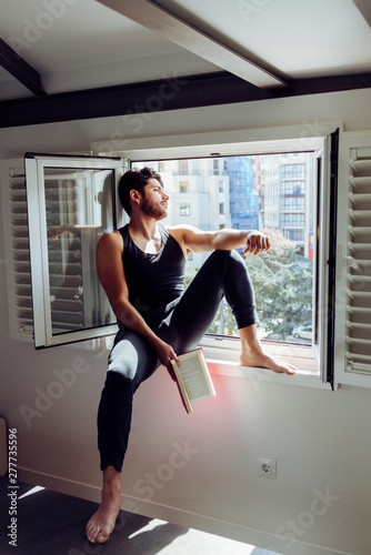 Side view of young serious handsome man in casual sleeveless shirt sitting on window and reading interesting book