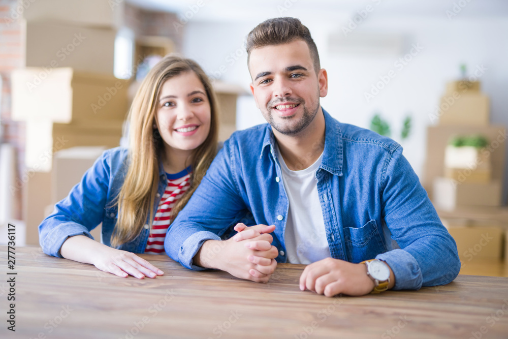 Young beautiful couple sitting on the table at home, hugging in love very happy for moving to new home with cardboard boxes behind them Young beautiful couple sitting on the table at home, hugging in love very happy for moving to new home with cardboard boxes behind them