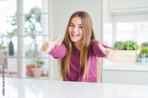 Free Download Beautiful young girl kid on white table approving doing positive gesture with hand, thumbs up smiling and happy for success. Looking at the camera, winner gesture. - GFXLoad