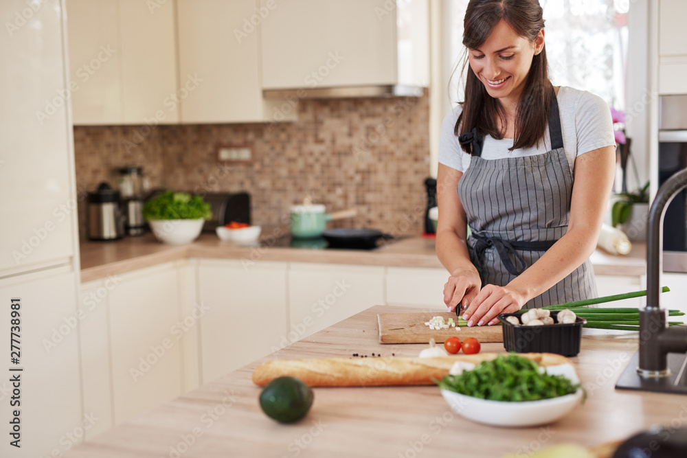 Smiling Caucasian female chef in apron chopping leek in kitchen and preparing dinner. Preparation of domestic food concept. Smiling Caucasian female chef in apron chopping leek in kitchen and preparing dinner. Preparation of domestic food concept.