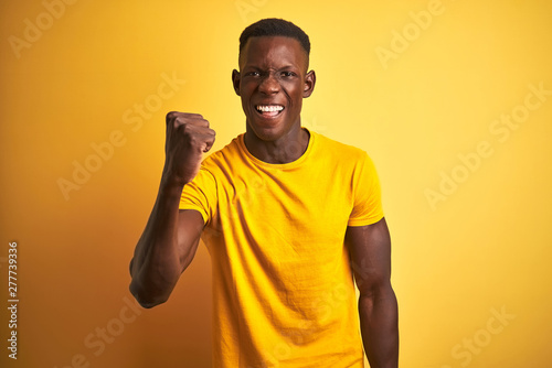 Young african american man wearing casual t-shirt standing over isolated yellow background angry and mad raising fist frustrated and furious while shouting with anger. Rage and aggressive concept.