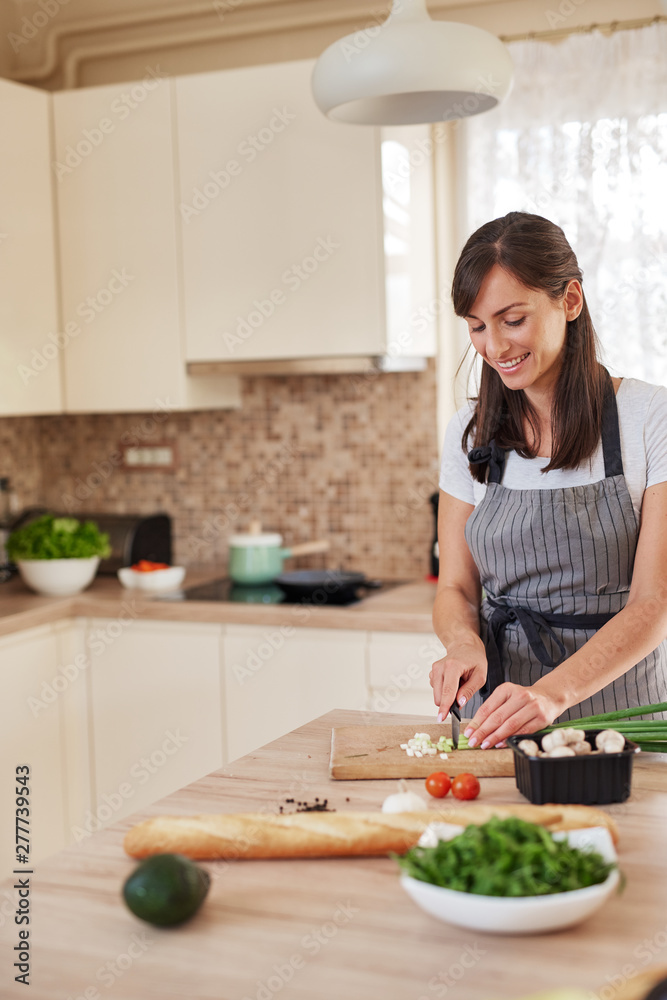 Smiling Caucasian female chef in apron chopping leek in kitchen and preparing dinner. Preparation of domestic food concept. Smiling Caucasian female chef in apron chopping leek in kitchen and preparing dinner. Preparation of domestic food concept.