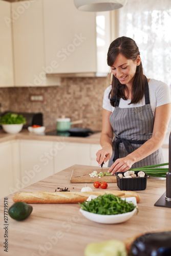 Smiling Caucasian female chef in apron chopping leek in kitchen and preparing dinner. Preparation of domestic food concept.
