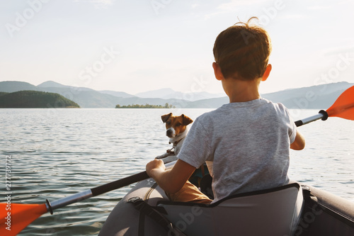 Happy boy with his dog Jack Russell Terrier paddling an inflatable kayak on the water mountain lake against the backdrop of beautiful orange sunset. Family sports vacation. Lens flare. Pet