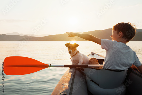 Happy boy with his dog Jack Russell Terrier paddling an inflatable kayak on the water mountain lake against the backdrop of beautiful orange sunset. Family sports vacation. Lens flare. Pet