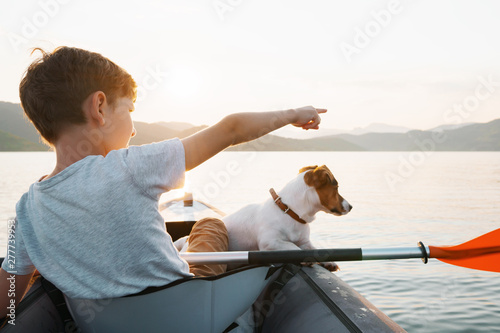 Happy boy with his dog Jack Russell Terrier paddling an inflatable kayak on the water mountain lake against the backdrop of beautiful orange sunset. Family sports vacation. Lens flare. Pet