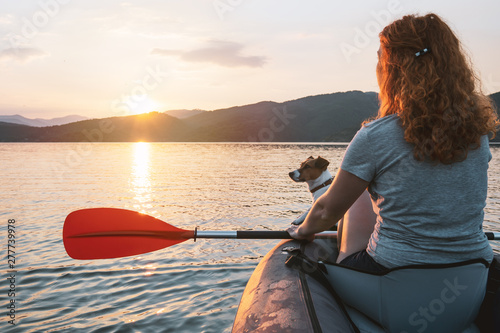 Happy young woman with her dog Jack Russell Terrier paddling on an inflatable kayak on the water of a large mountain lake against a beautiful orange sunset. Family Sports Weekend