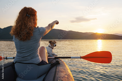 Happy young woman with her dog Jack Russell Terrier paddling on an inflatable kayak on the water of a large mountain lake against a beautiful orange sunset. Family Sports Weekend