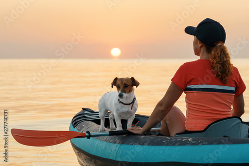 Red-haired young woman is rowing on an inflatable kayak by the sea with a dog Jack Russell Terrier on a background of pink sunrise in beautiful nature. Great disk of the rising sun. Sun rays. Sport