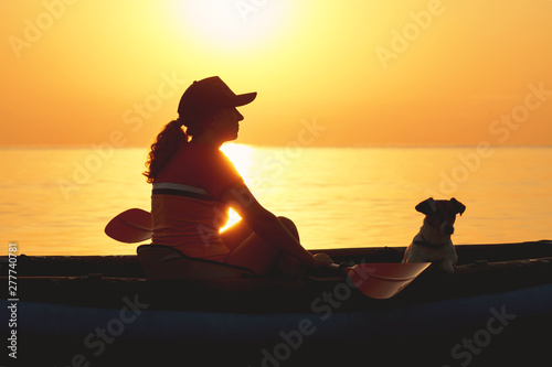 Red-haired young woman is rowing on an inflatable kayak by the sea with a dog Jack Russell Terrier on a background of pink sunrise in beautiful nature. Great disk of the rising sun. Sun rays. Sport