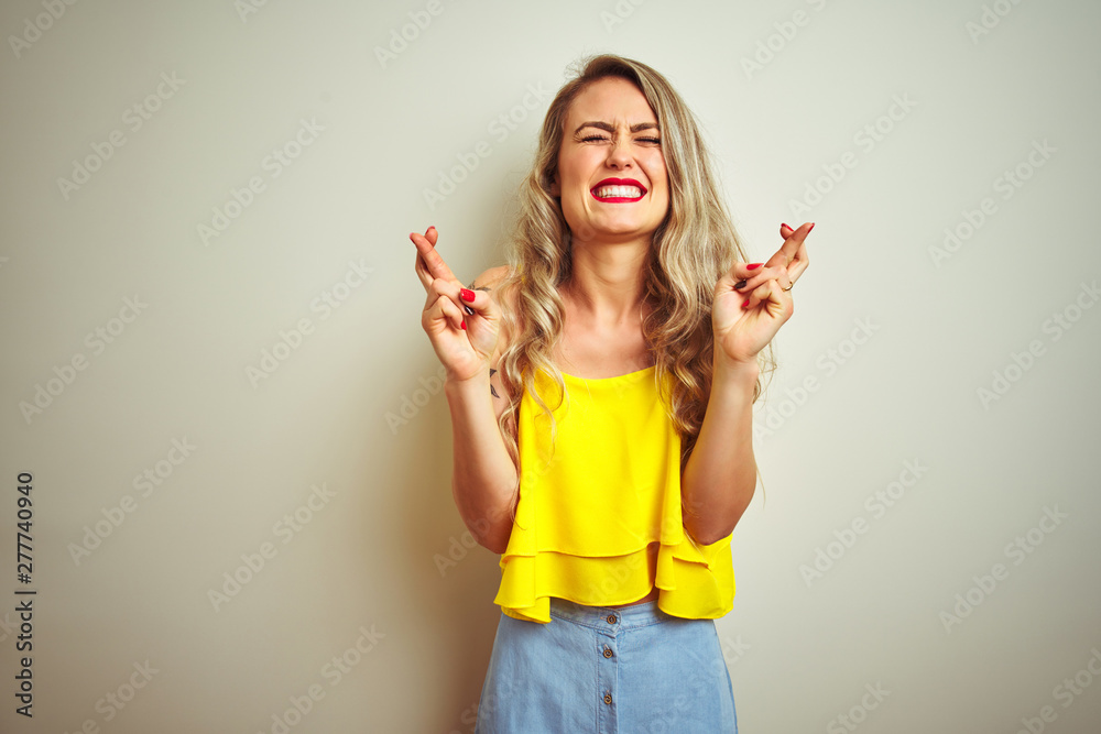 Young beautiful woman wearing yellow t-shirt standing over white isolated background gesturing finger crossed smiling with hope and eyes closed. Luck and superstitious concept. Young beautiful woman wearing yellow t-shirt standing over white isolated background gesturing finger crossed smiling with hope and eyes closed. Luck and superstitious concept.